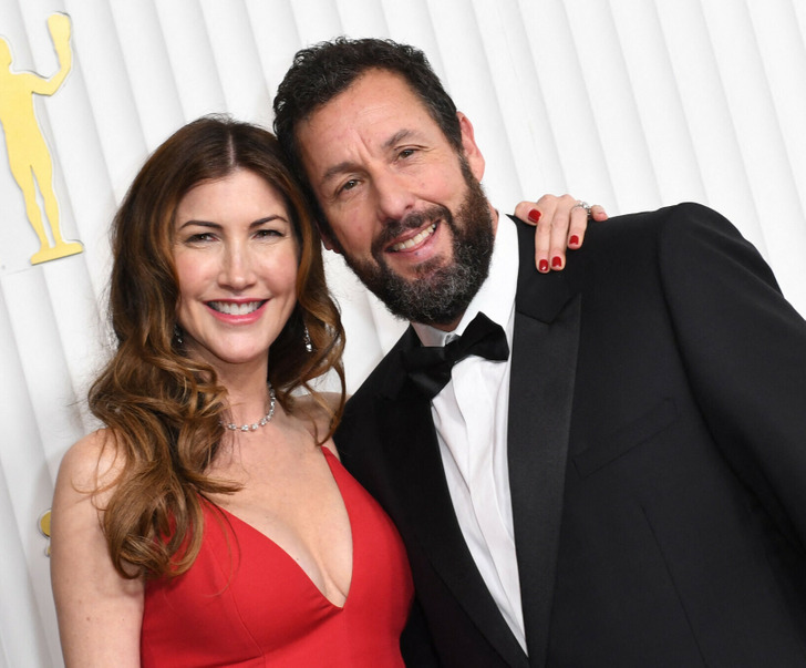 Adam Sandler wearing a black suit and bow tie with his wife on the red carpet.