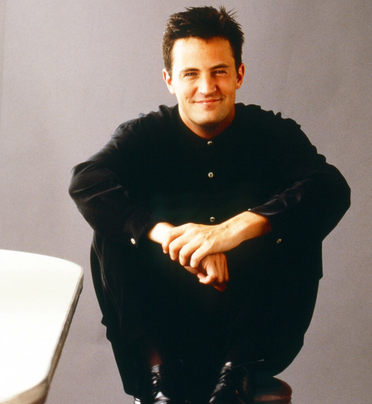 Matthew Perry posing for the camera sitting on bar stools wearing all black.