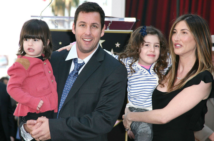 Adam Sandler wearing suit and tie holding one of his daughters together with his wife who is holding the other daughter.