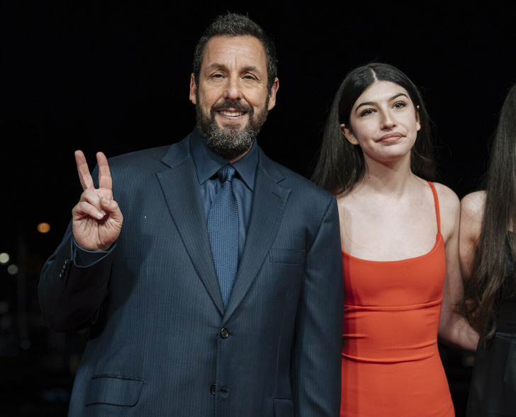 Adam Sandler wearing a dark blue suit and tie with his daughter who is wearing a red dress.